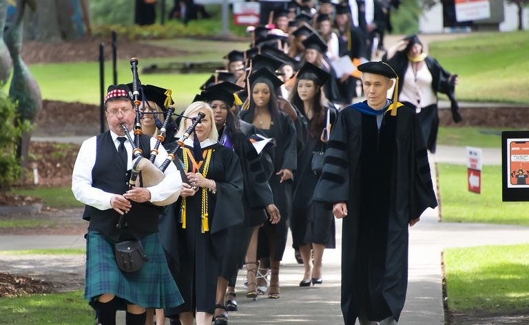 A Processional Of College Students Wearing Black Caps And Gowns Walks Toward The Auburn University At Montgomery Athletics Complex.