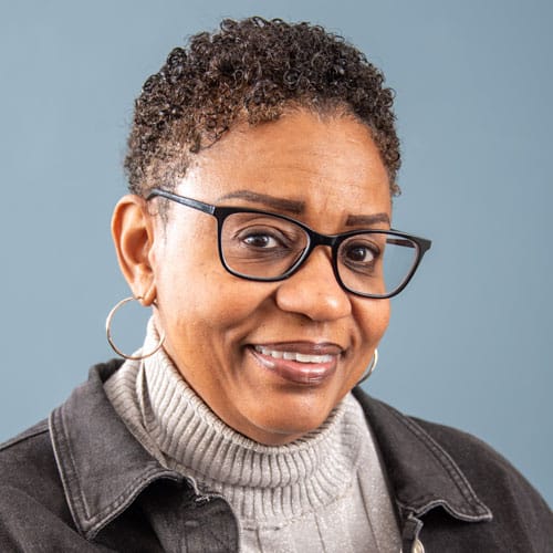 A person wearing glasses and a turtleneck sweater smiles against a plain backdrop.
