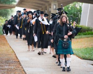 A processional of college students wearing black caps and gowns walks toward the Auburn University at Montgomery Athletics Complex.
