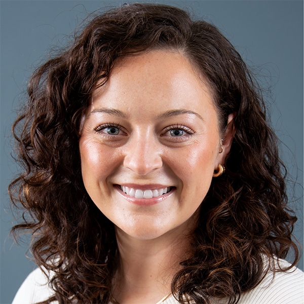 A smiling woman with curly hair and gold earrings against a gray background.