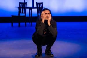 A Person Squatting On Stage Under Blue Lighting, Holding Face In Hands, With Chairs In The Background.