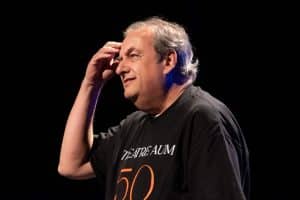 A Man In A Black Shirt With THEATRE AUM 50 Stands On Stage, Touching His Forehead Thoughtfully.