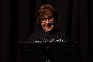 A Woman Wearing Glasses And A Theatre AUM Shirt Stands In Front Of A Lectern On A Dark Stage.