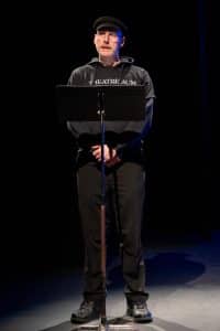 A Person Wearing A Theatre AUM Shirt And Hat Stands At A Podium On Stage, Under A Spotlight.