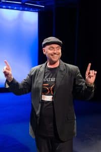 A Man Wearing A Black Hat And Jacket Gestures On Stage Under Blue Lighting At A Theater Event.