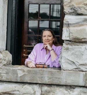 A woman in a purple shirt leans on a stone window ledge with a contemplative expression.