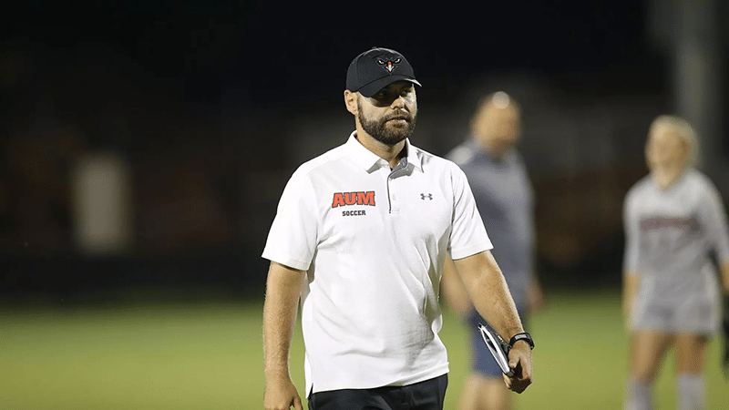 AUM soccer coach on the field during a night game, wearing a cap and holding a clipboard.