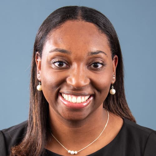 A woman smiling against a blue background, wearing pearl earrings and necklace.