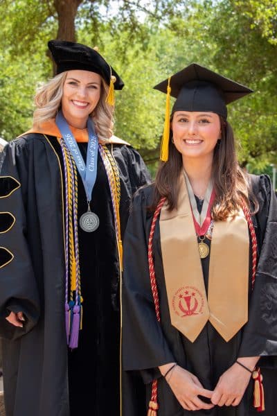 AUM faculty member Ashley D'Elia and her stepdaughter student Kennedy D'Elia smiling in regalia outdoors on campus.