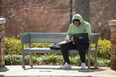 Auburn University at Montgomery student studies on campus bench with laptop and notebook outdoors.