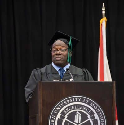 AUM commencement speaker Pookie Wilson in cap and gown at podium with Auburn University at Montgomery seal and Alabama flag.